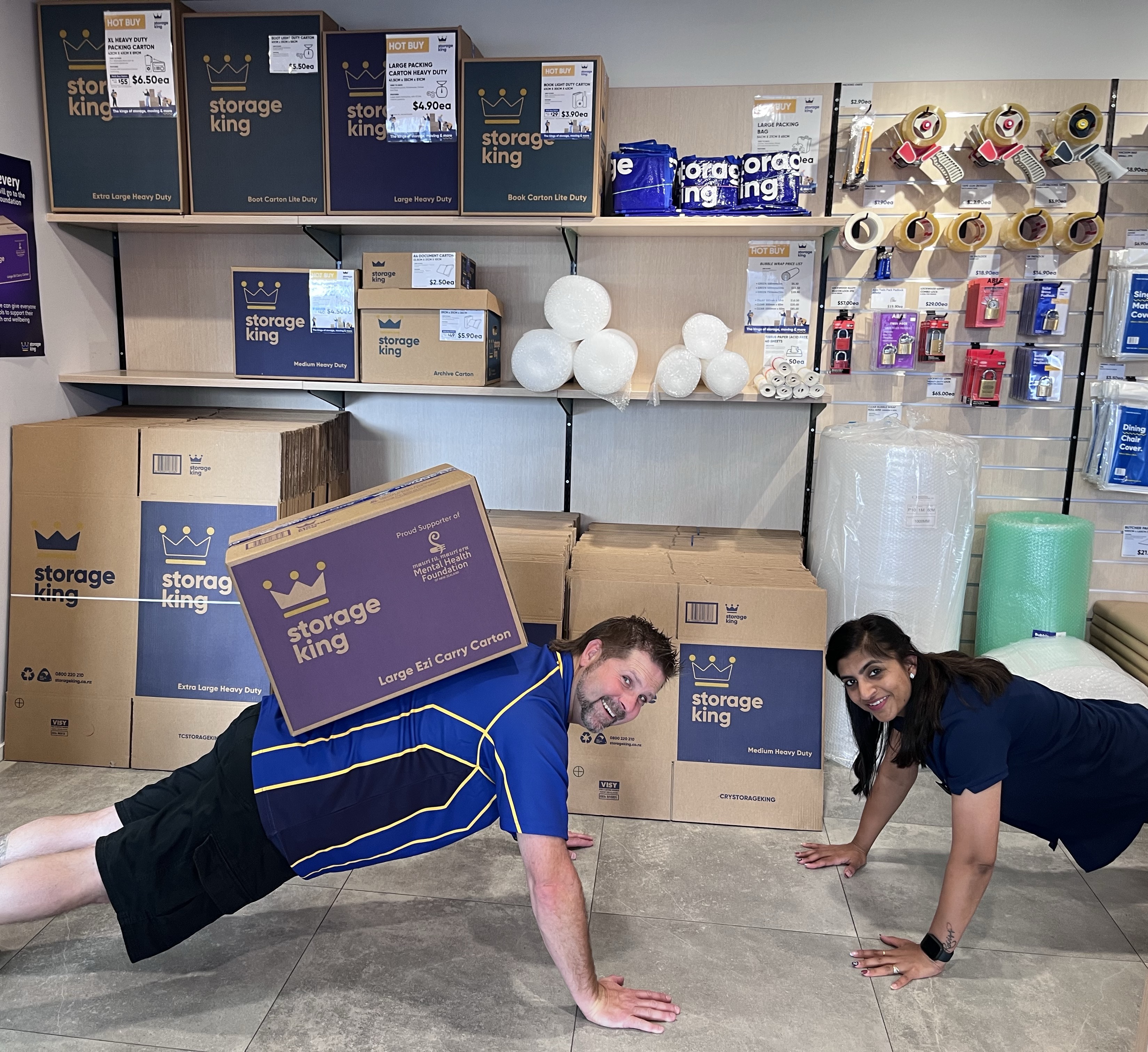 Two people smiling for the camera as they do push-ups in a Storage King warehouse, one of them has a big Storage King box on their back.