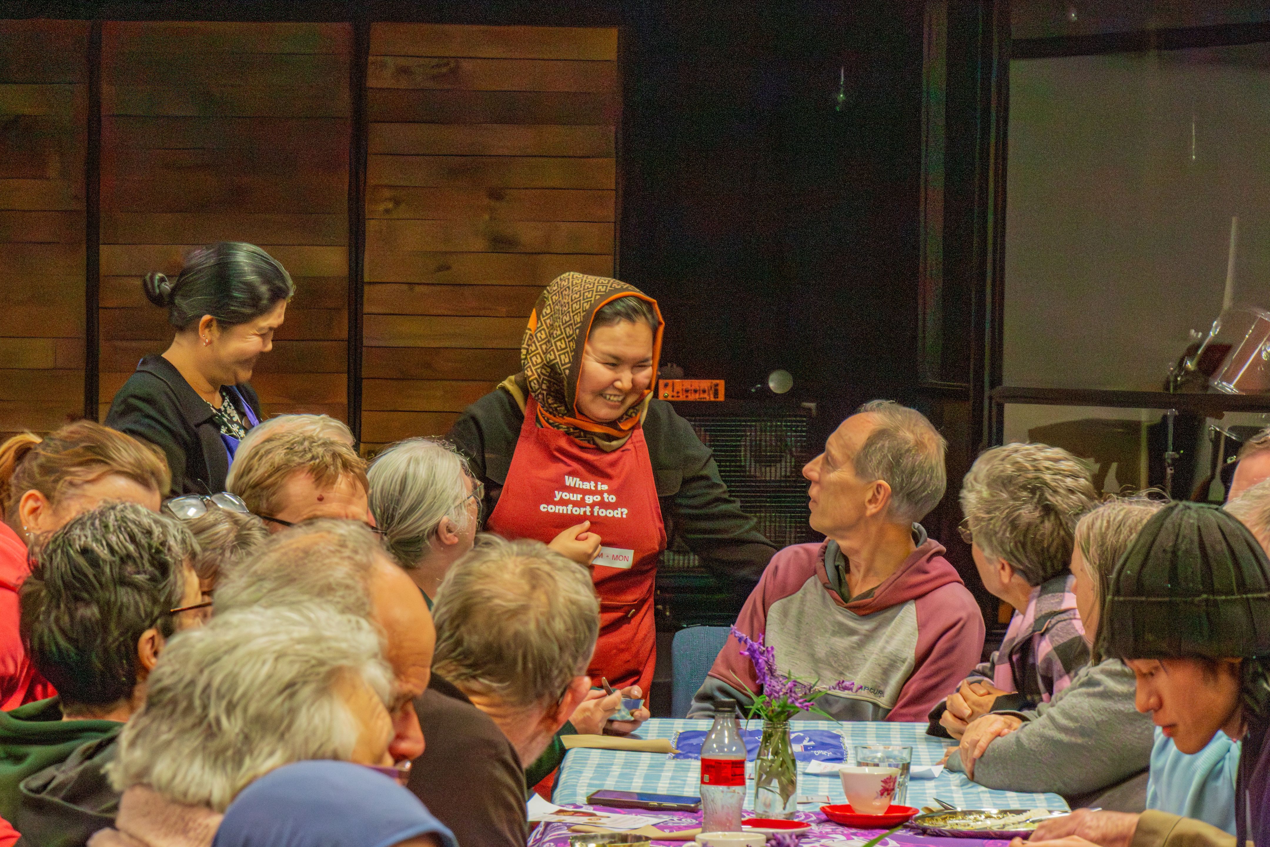 Volunteer in a red apron speaks with guests seated around long tables during a busy community shared meal gathering.