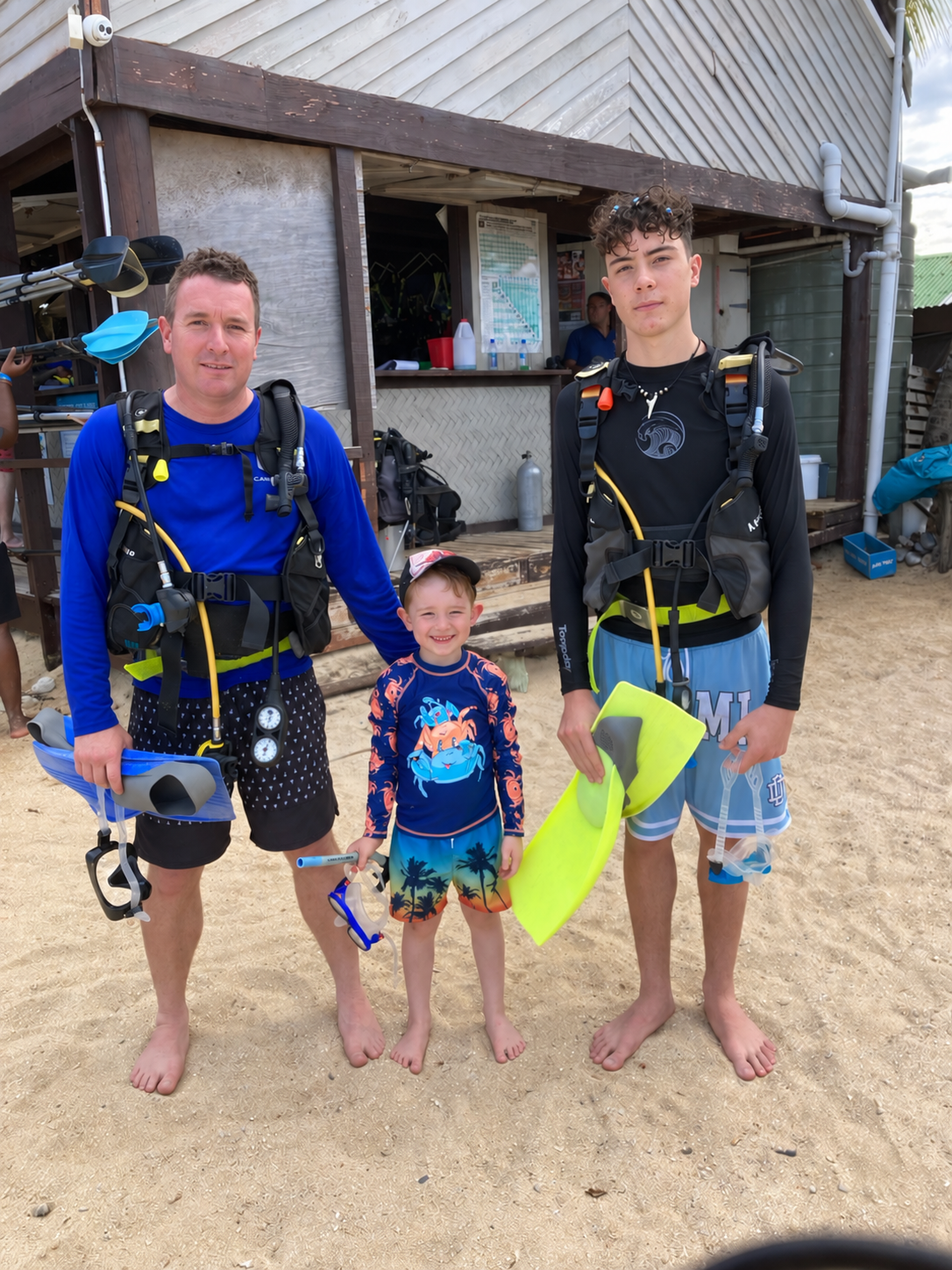 Three people in aquatic gear, carrying flippers and goggles, stood barefoot in the sand.