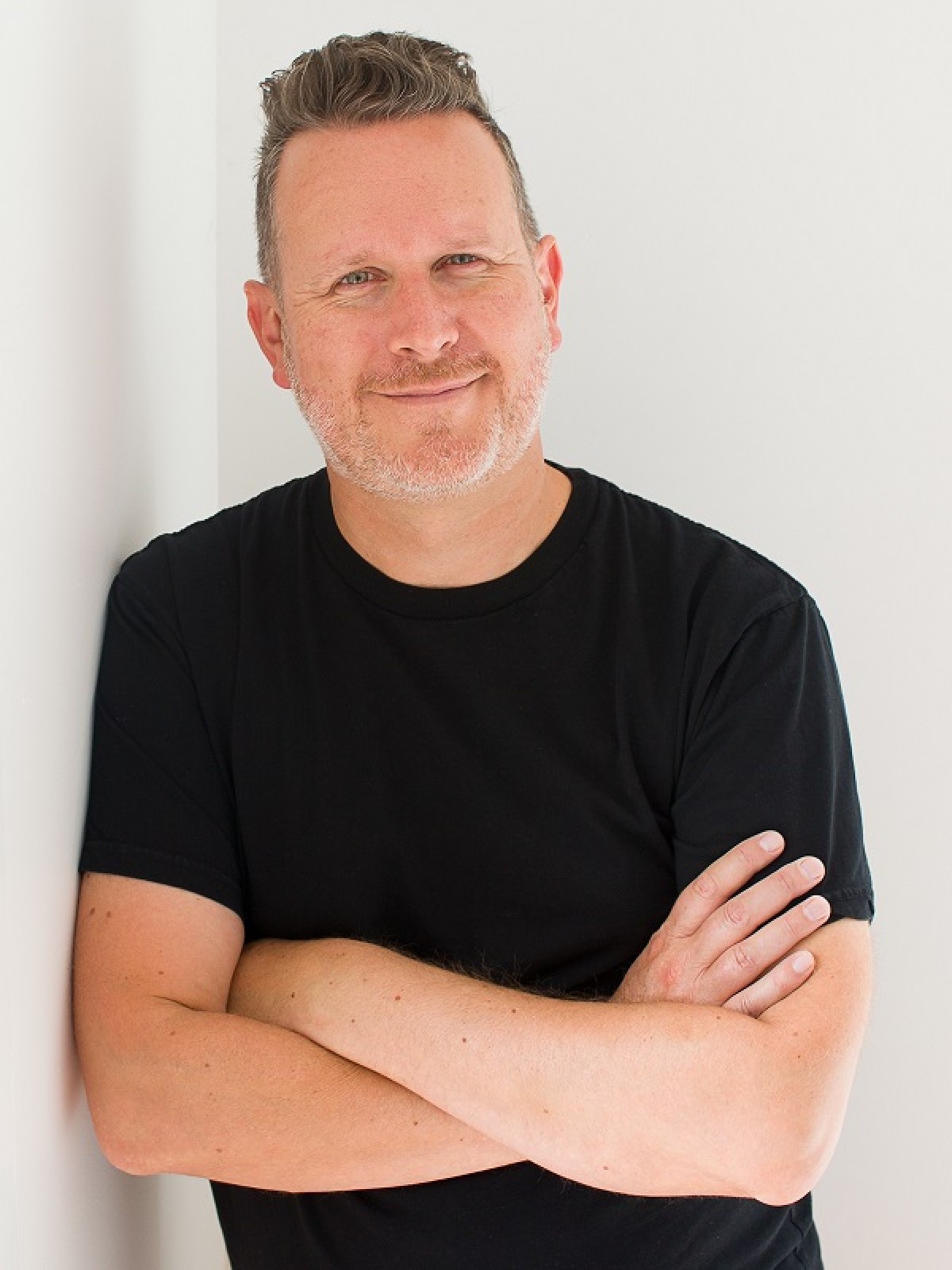 Man in a black T-shirt, friendly smile, arms crossed, leans against a wall to his right.