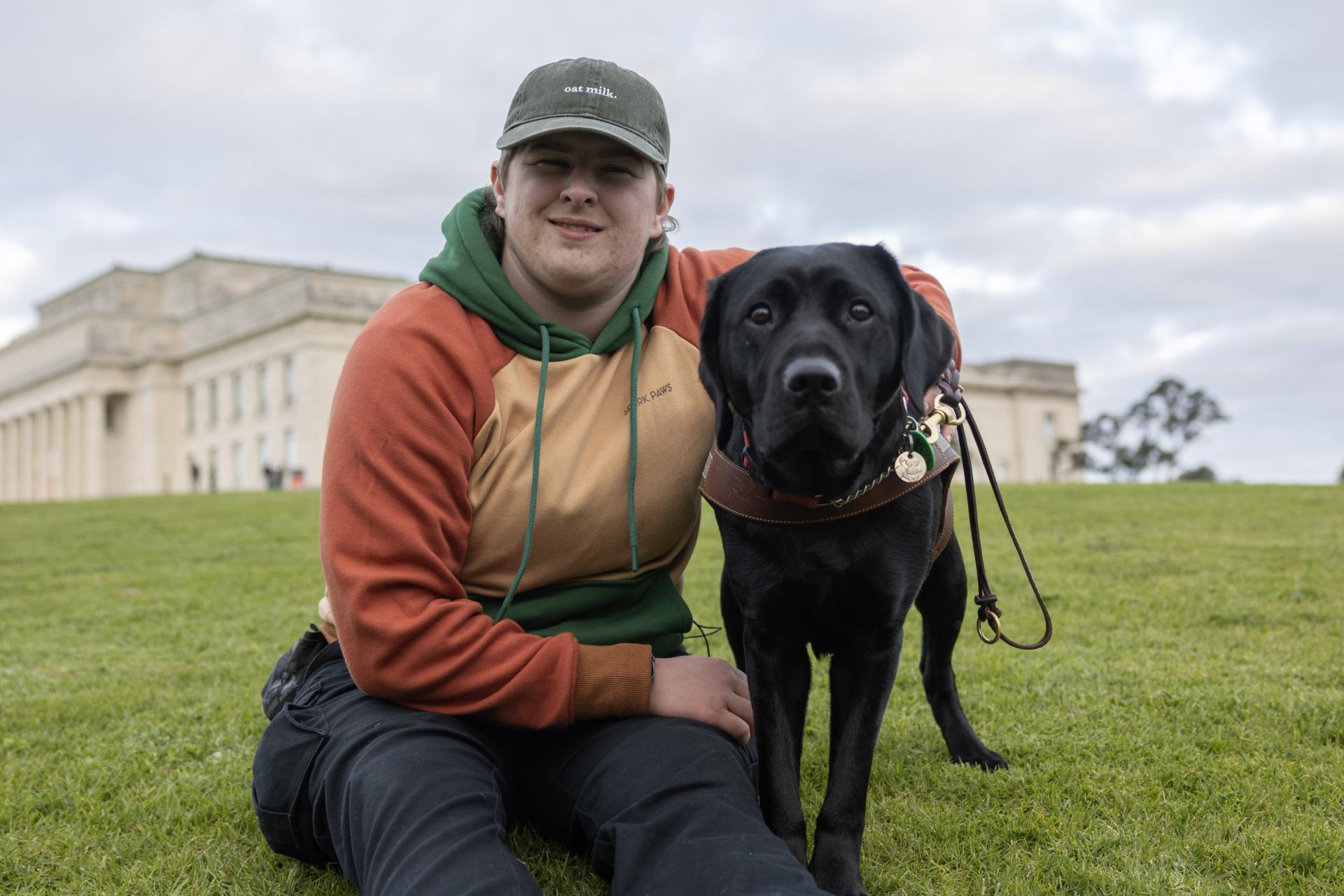 Person in a baseball cap and colourful hoodie poses with their black Labrador guide dog.