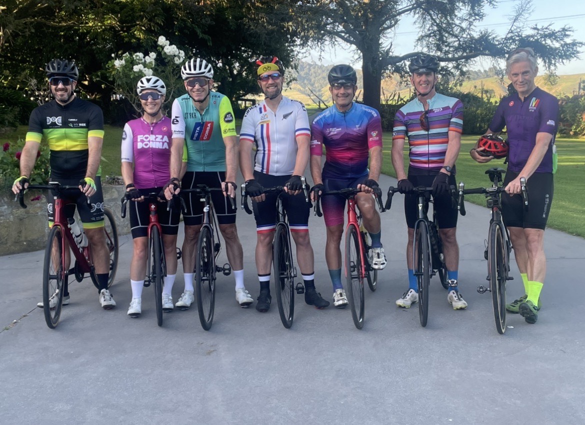 Seven cyclists in colourful cycling gear and helmets, smiling for the camera.
