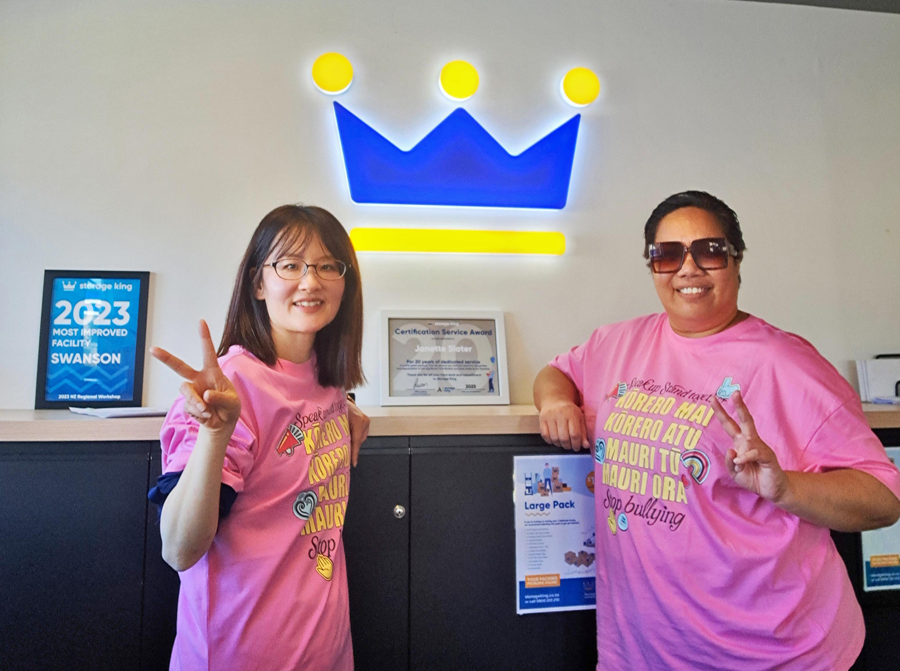 Two people in front of a bright blue King Storage logo, posing in their Pink Shirt Day T-shirts, giving us a v for victory sign.