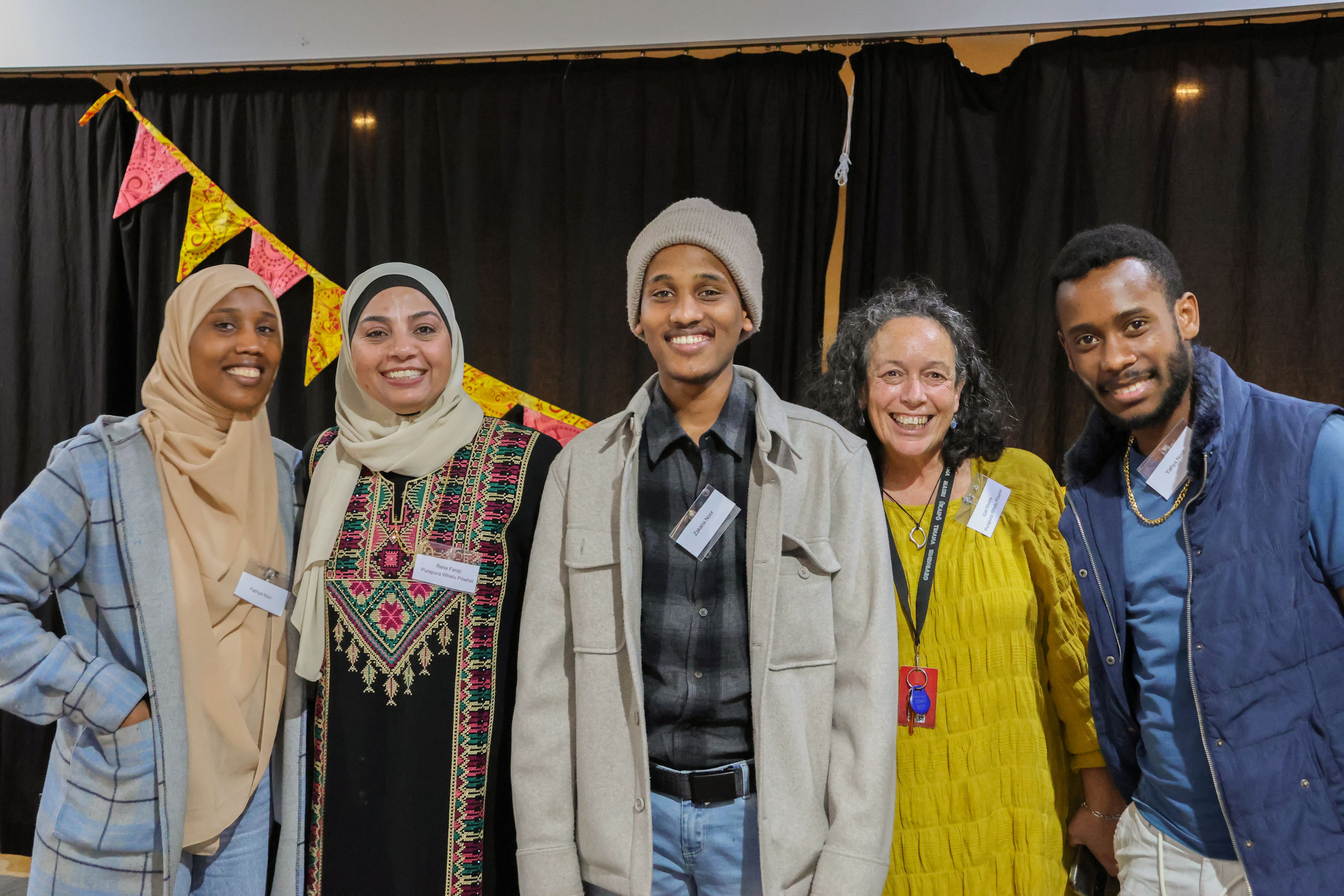 Group of five attendees stand in front of colourful bunting at a community gathering, wearing name badges and smiling for the camera.