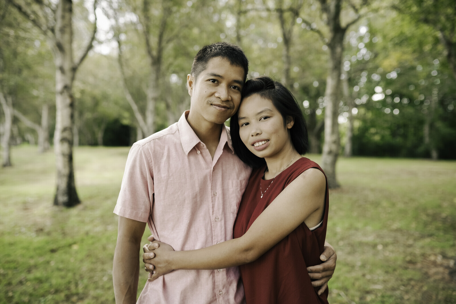 Two people embracing in a park surrounded by trees and greenery.