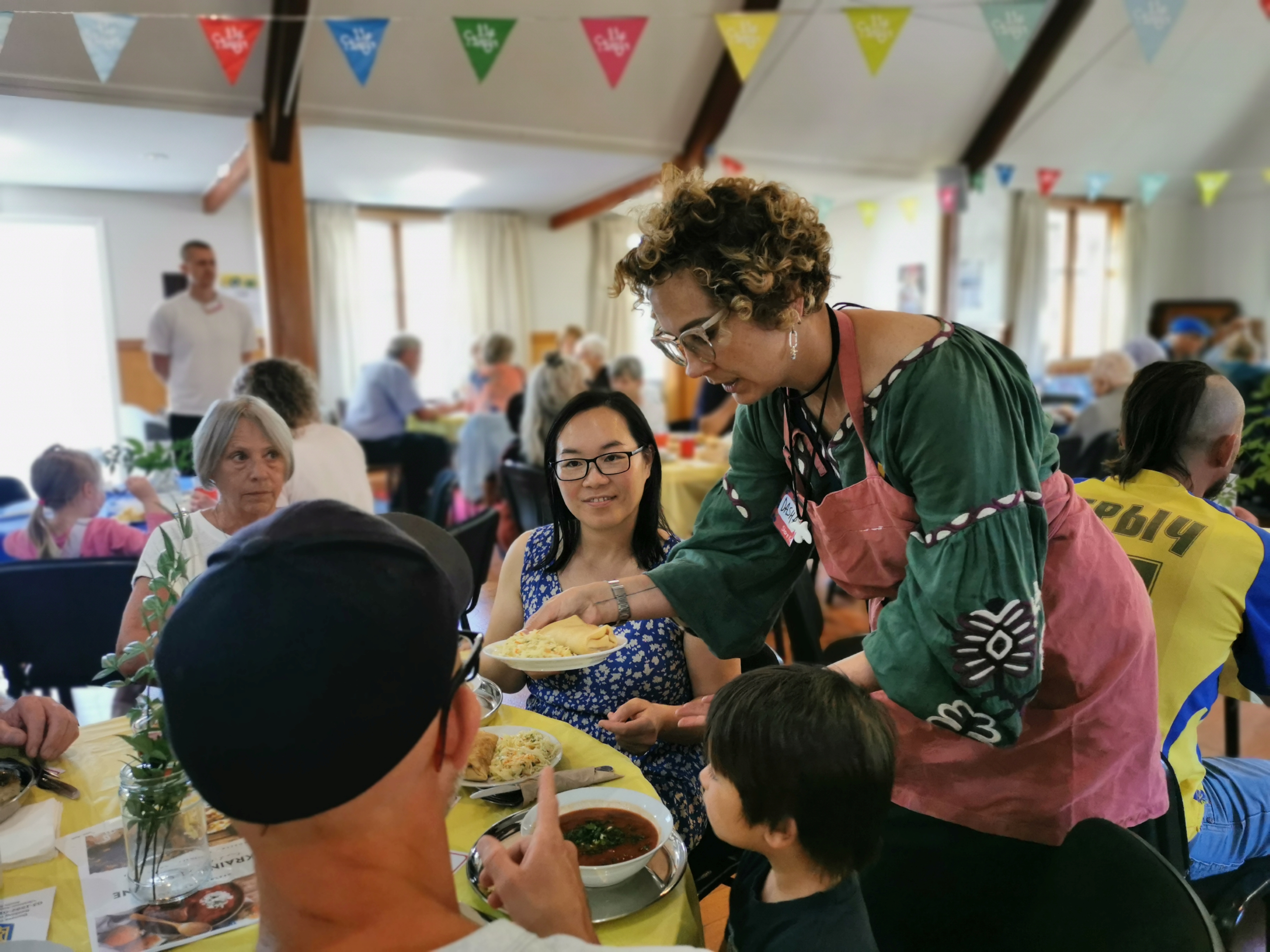 Person in an apron serves a plate of food to attendees seated around a table at a busy community meal in a decorated hall.