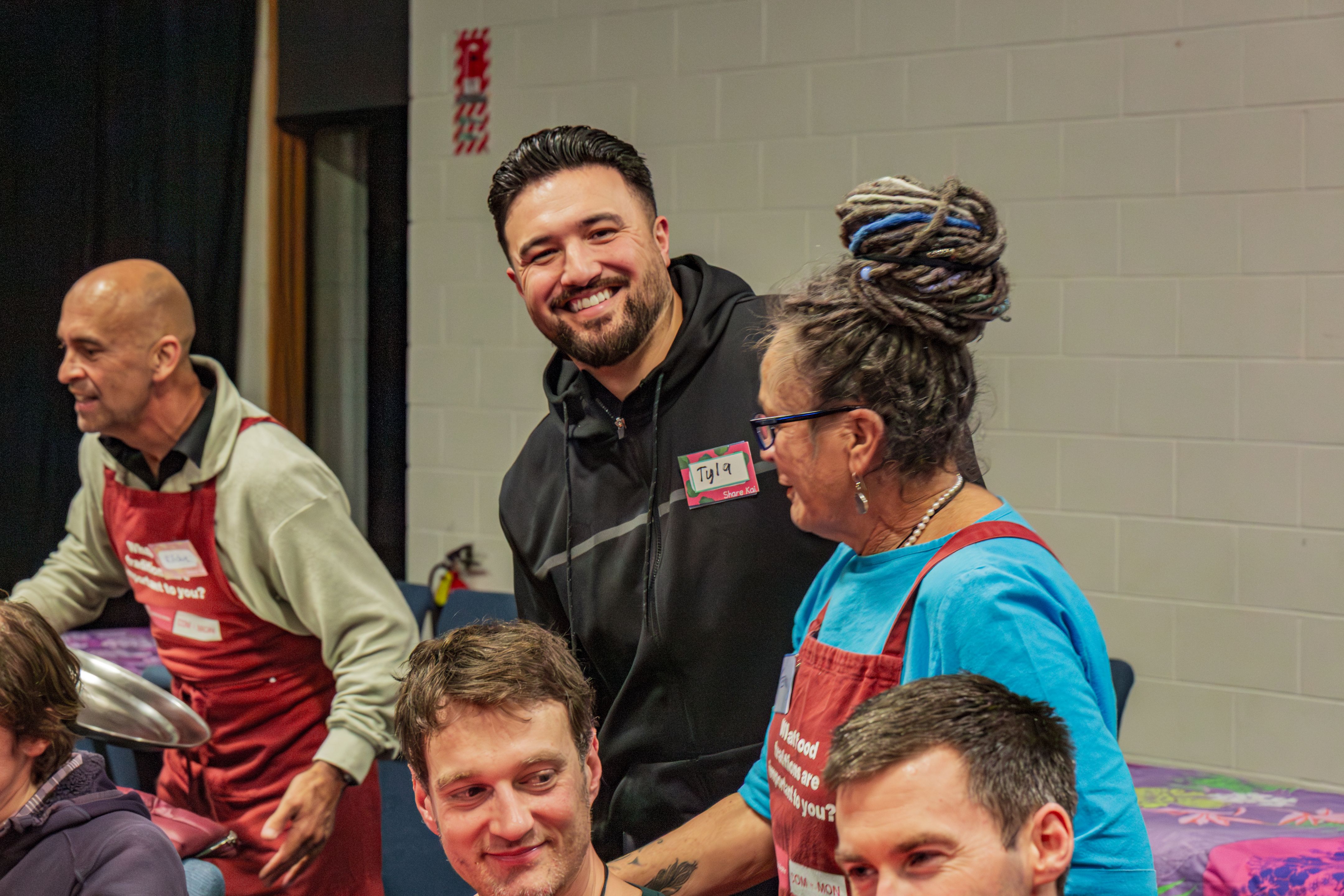 Volunteers in red aprons and community attendees gather around shared tables in a hall, chatting and serving food during a community meal event.