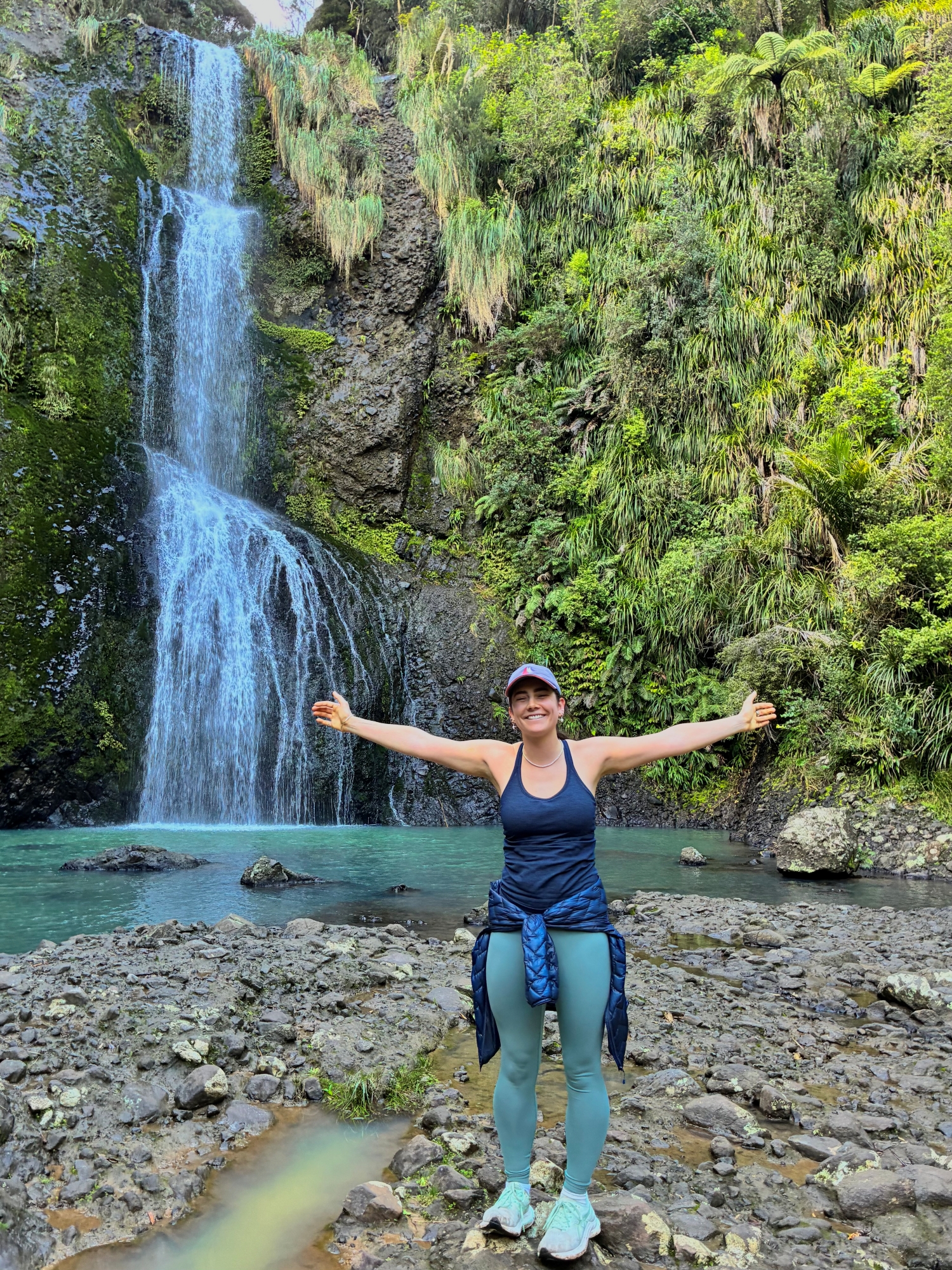 Woman in activewear smiles and poses with her arms wide open in front of a tall waterfall.