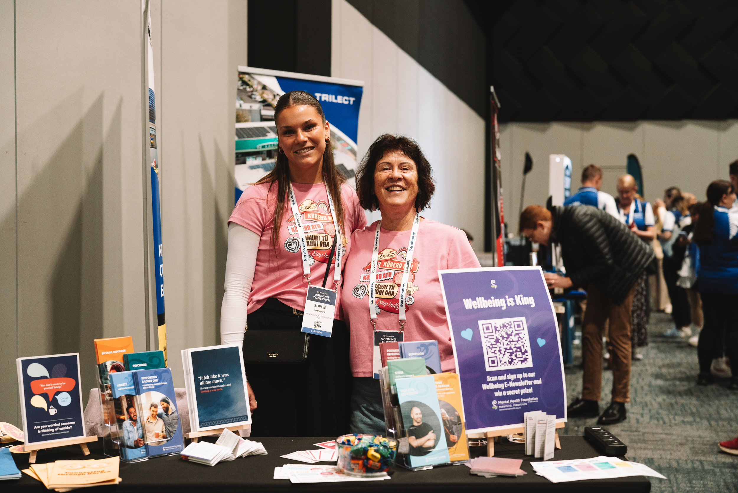 Two hosts at an exhibition stand, in Pink Shirt Day T-shirts, smiling for the camera. In front of them is a poster that says Wellbeing is King.