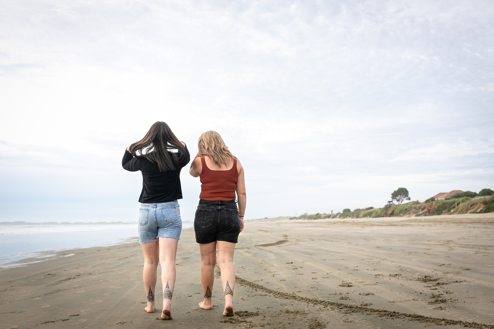 Two people walk barefoot along a quiet beach under an overcast sky.