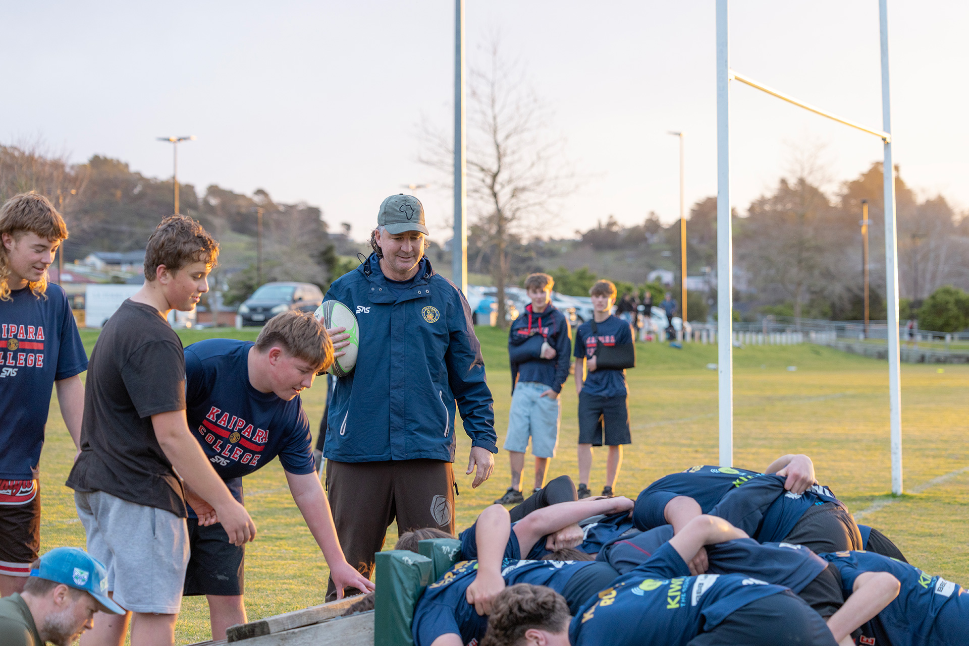 Rugby players practise a scrum under a coach’s guidance on a sports field at sunset.
