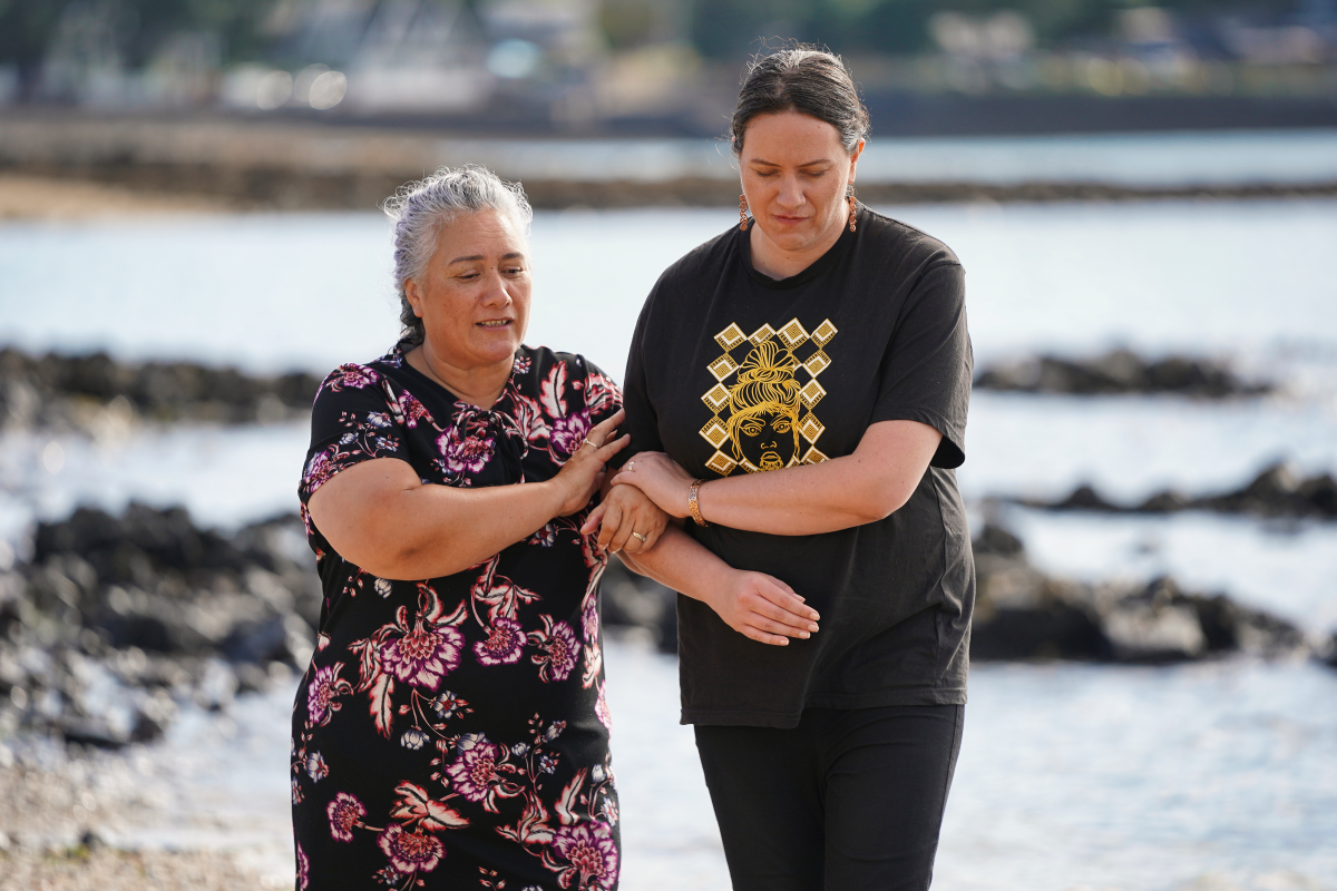 Two people walk together along a rocky shoreline, with one person gently supporting the other's arm.