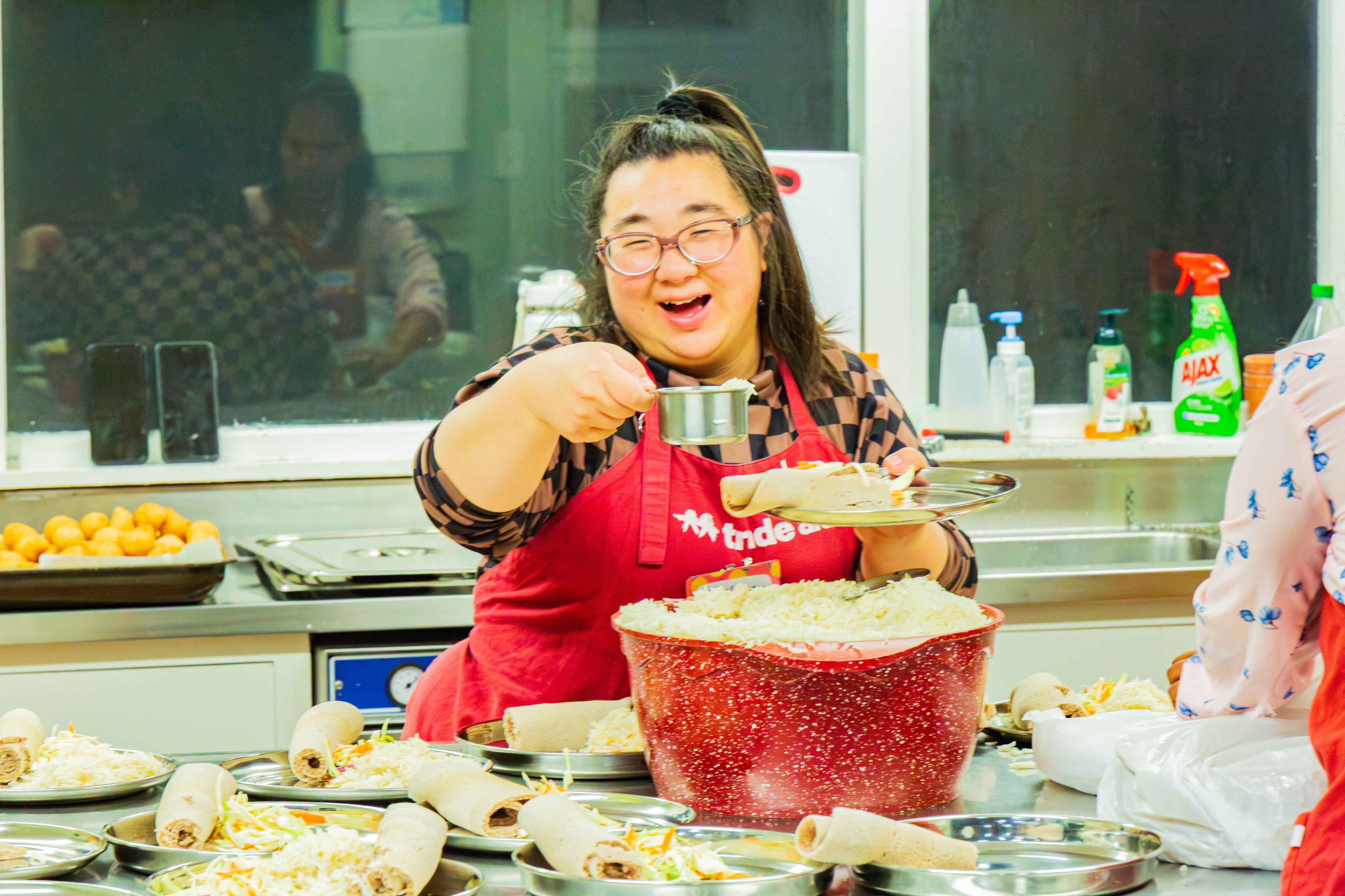 Volunteer in a kitchen wearing a red apron serves food onto plates beside a large bowl of noodles during a community meal preparation.