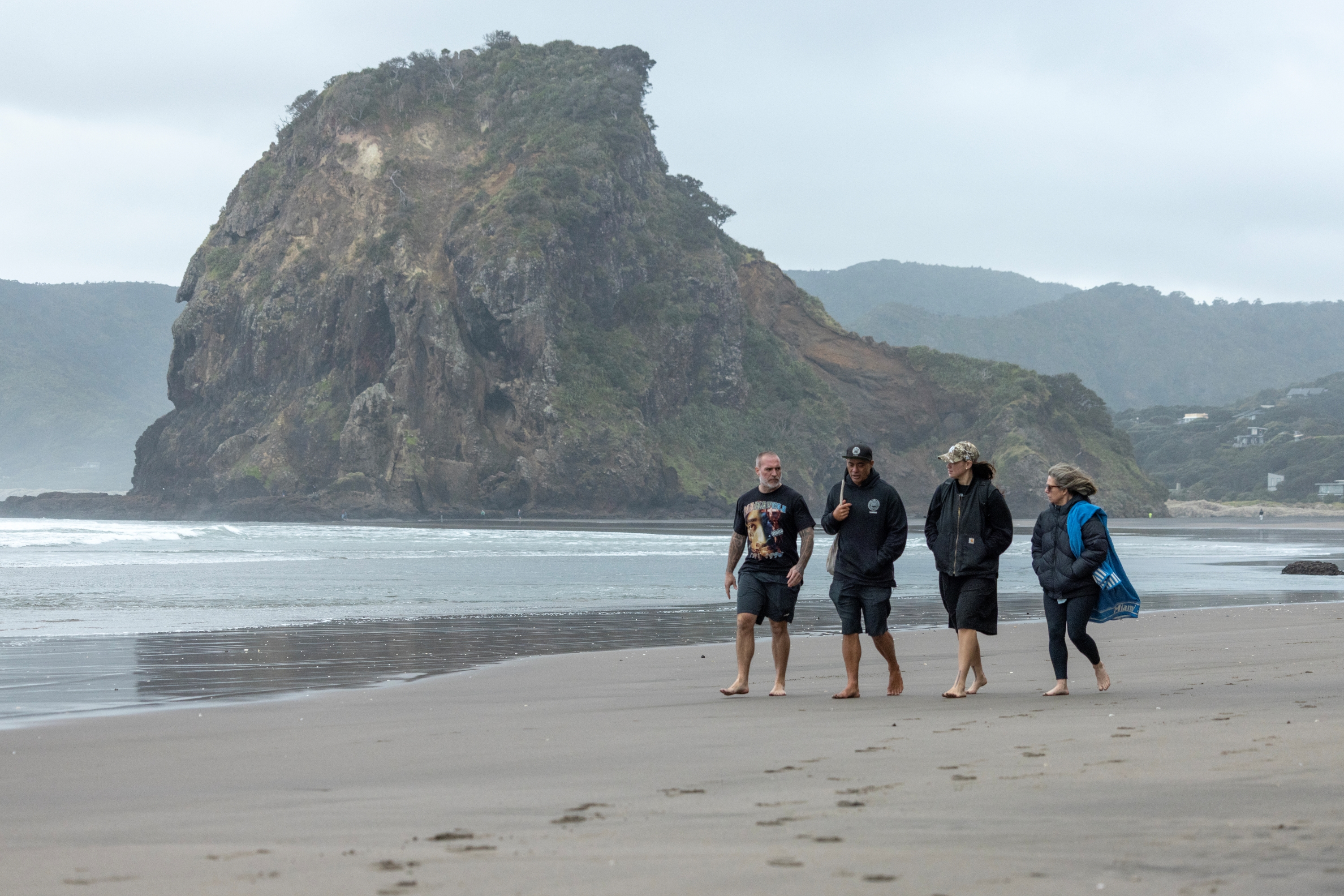 Four people in conversation walk along the shore of a beach, it appears to be windy and cold.